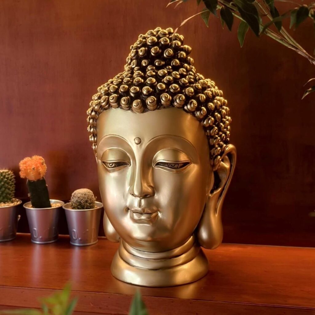 A golden Buddha head statue, with detailed features. Plants are also visible.