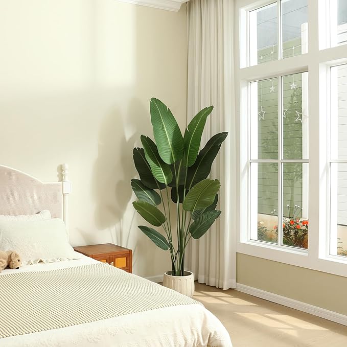 A bedroom with a large plant, bed, and window. Cream walls and light colored bedding.