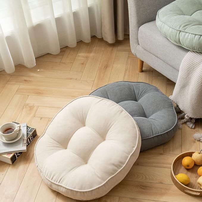 Two round floor cushions, one light beige, one gray, are on a light brown wooden floor. A couch is in the background.