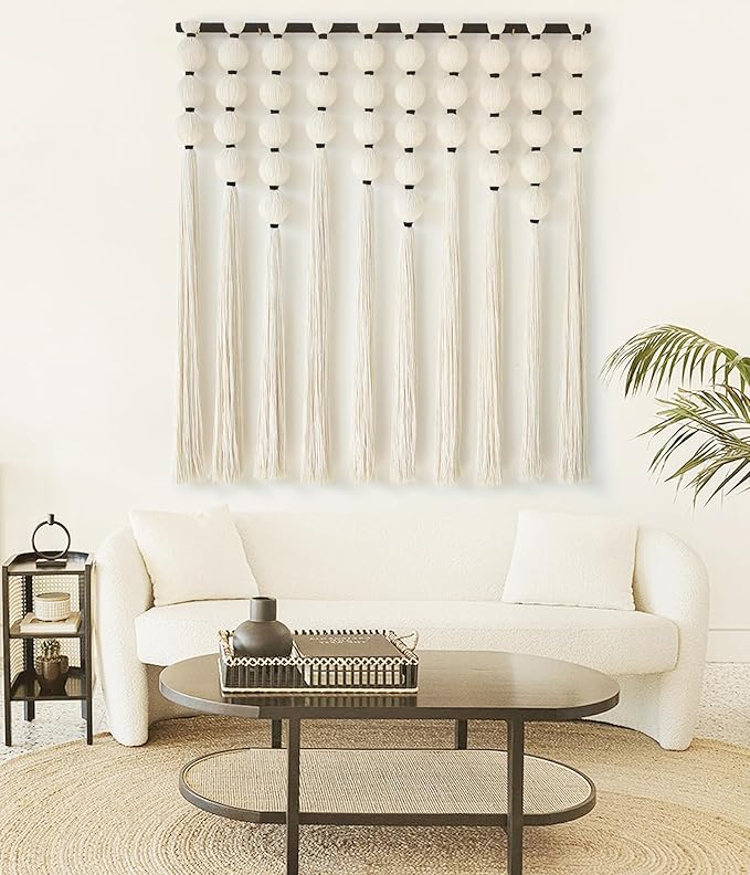 A cream colored wall hanging with tassels and small balls, a white couch, and a coffee table.